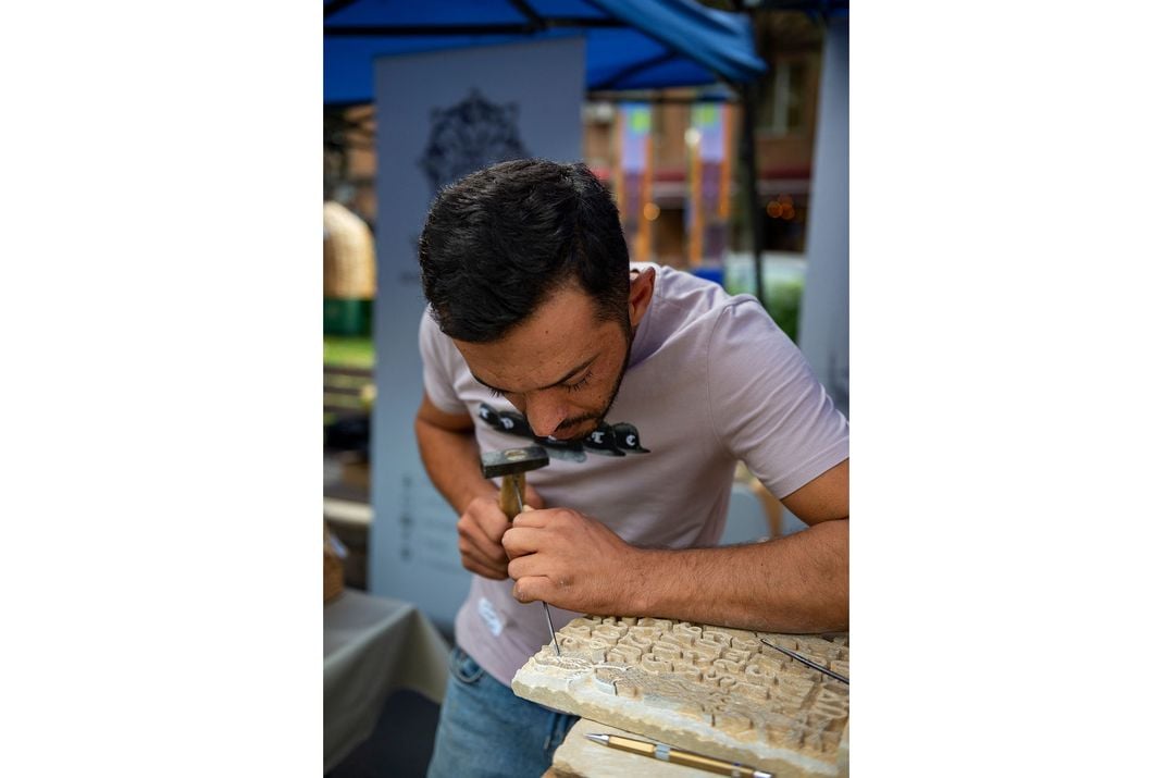 A young man wearing a blue shirt bends over a table to carve a piece of stone.