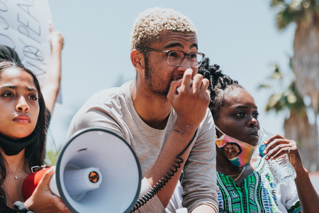 Three young activists are outside at a protest – looking off into the distance. They all appear to be African American. One is holding a bullhorn and has a determined look on her face. Another is holding the bullhorn’s microphone and speaking into it.