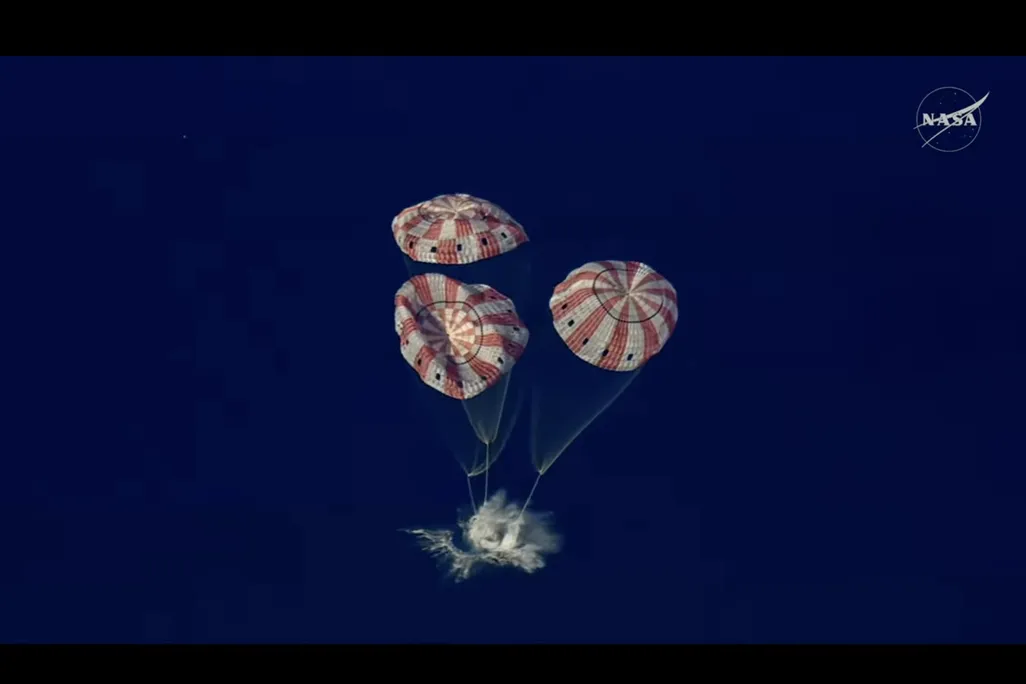 spacecraft splashing down in the ocean with three parachutes above