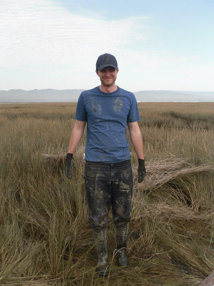 James Holmquist stands in a grassy wetland, wearing field pants, boots and a blue T-shirt caked with mud