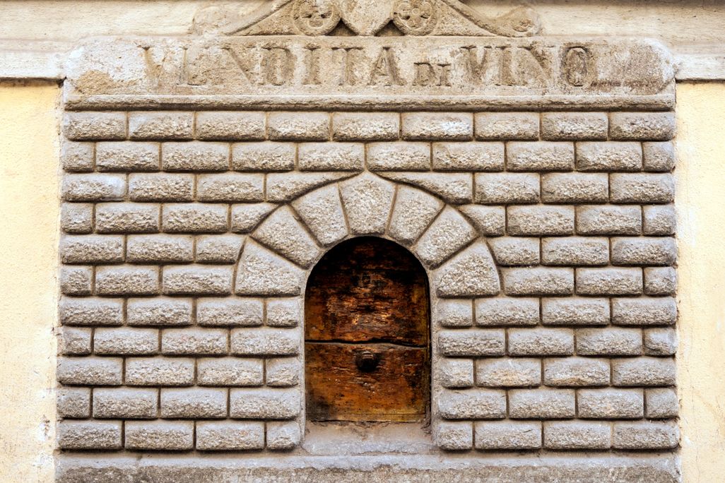 A small door cut into the side of a house. The door has engraved stone around its entrance, made to look like a tiny building facade. It bears an inscription that has worn away, but includes the word "VINO"