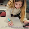 A student leans over a table, writing on a large poster board with a marker during a classroom activity. Surrounding the workspace are colorful pens, papers, and notes, while additional tables, chairs, and a kitchen area with cabinets and a counter appear
