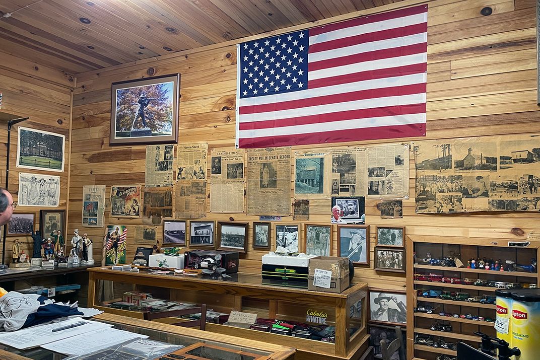 A wood-paneled interior corner of a store, with an American flag, newspaper clippings, and framed photos on the wall.