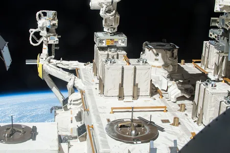 A robotic arm places a container with three panels of bacteria outside the International Space Station.