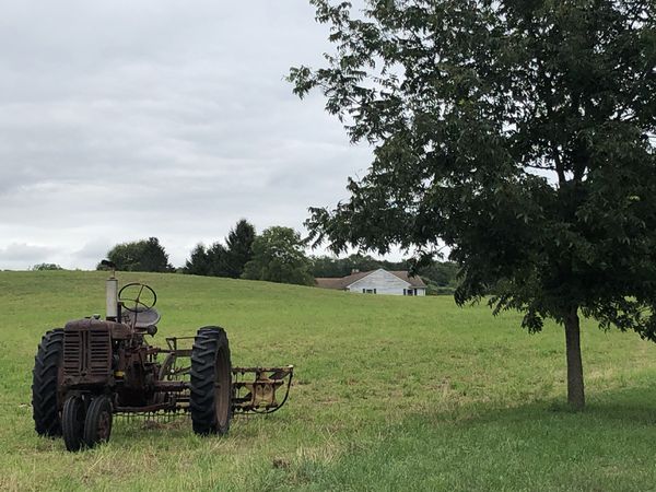 Old farm tractor in a summertime country field setting with an old house in the distance thumbnail