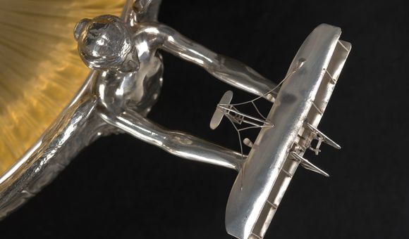 Overhead detail view of a trophy showing a silver angel extending her arms holding a vintage airplane.