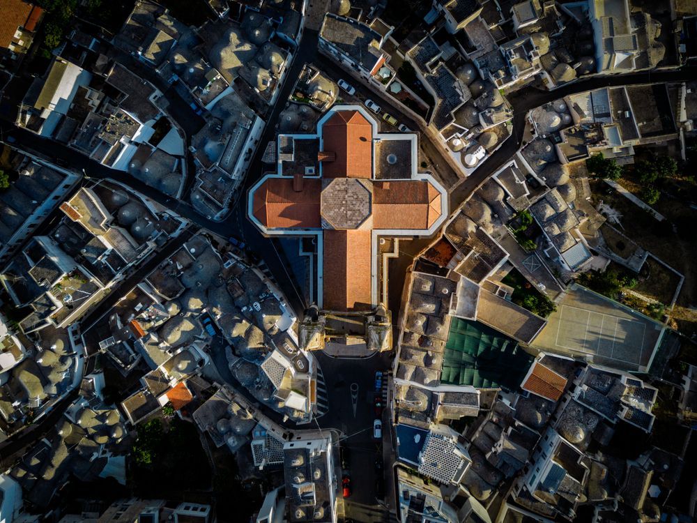 An aerial view of Alberobello, capturing the church rising above the landscape and the iconic Trulli surrounding it, forming a unique pattern of conical roofs.