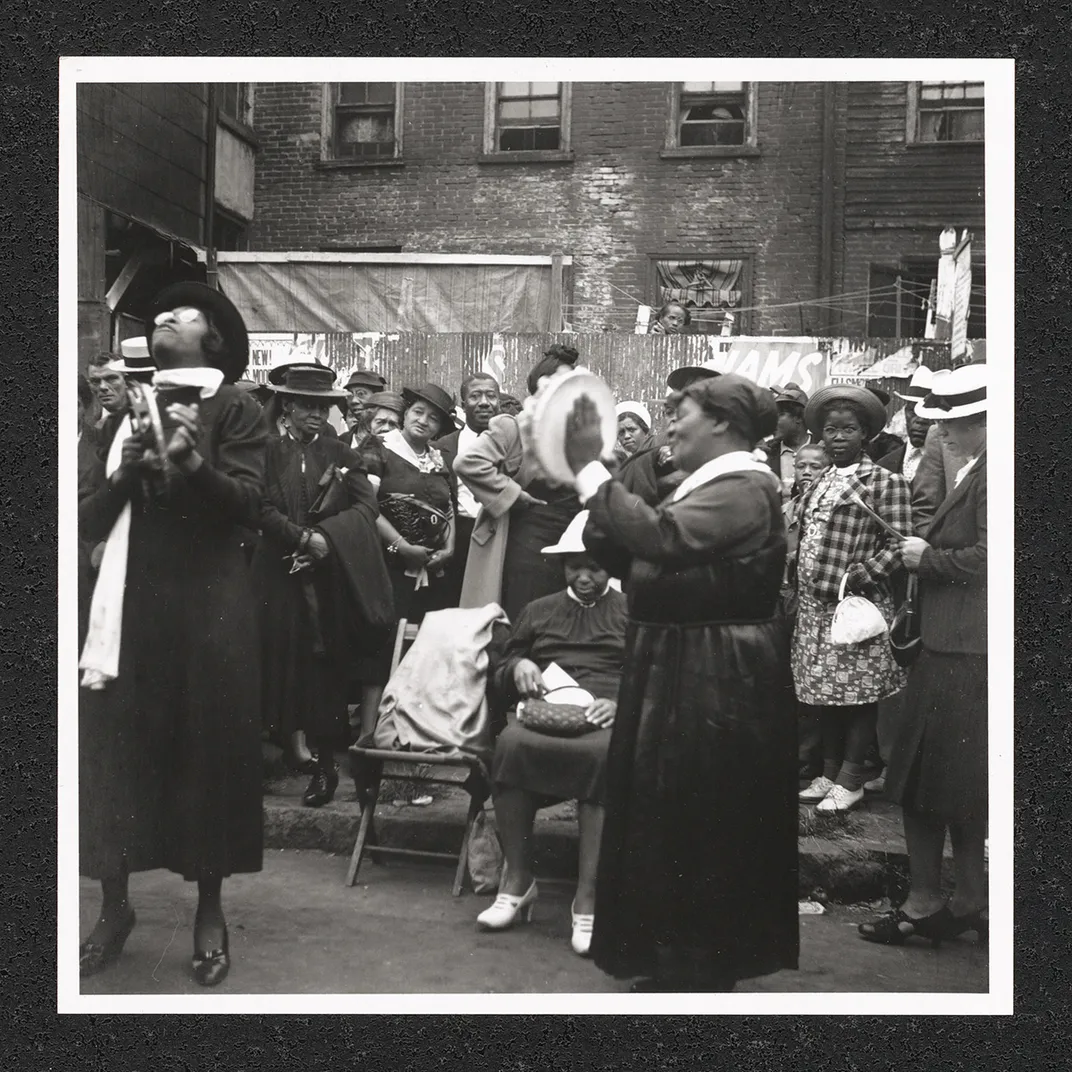 Two women standing in the street, playing tambourines in front of a crowd. Black-and-white photo.