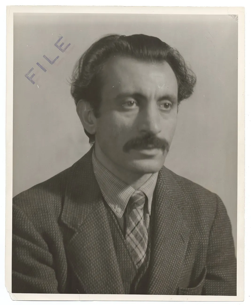 A black and white photograph of a mustachioed man with dark hair, in suit and tie, looking into the distance with a serious expression