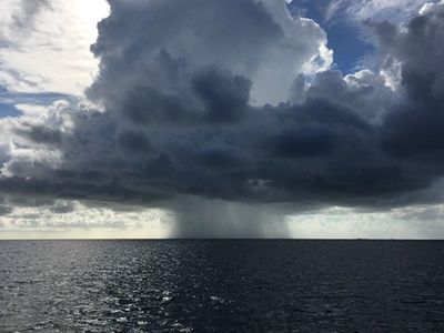 Rain falls from a small group of clouds over the open ocean as the sun shines behind the clouds.