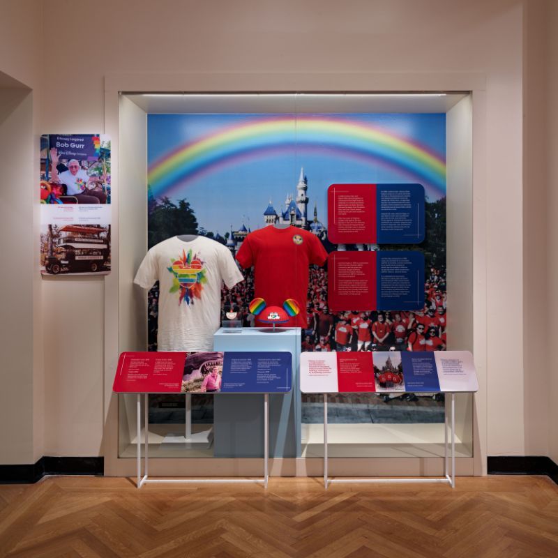 Exhibit features pride themed Disney merchandise like shirts and Mickey ears. There is a large mural of the park Castle with a rainbow overhead.