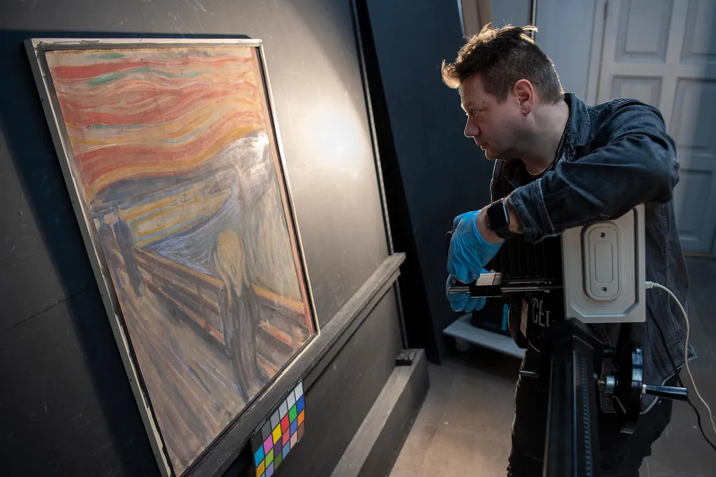 A white man in a blue shirt and gloves holds a camera in front of the canvas, which is about as tall as his torso