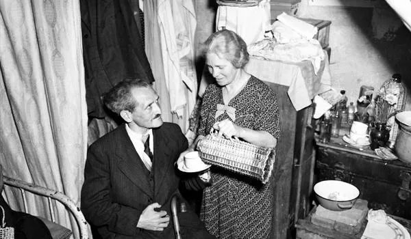 A white woman in a dress pours tea for a seated man in a suit, in a small home crowded with items (mobile)