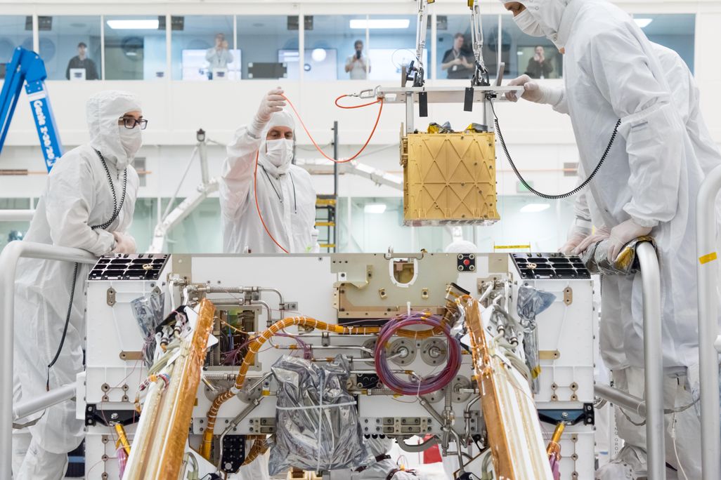 People in lab coats lower a gold box containing an oxygen-producing device into a Mars rover
