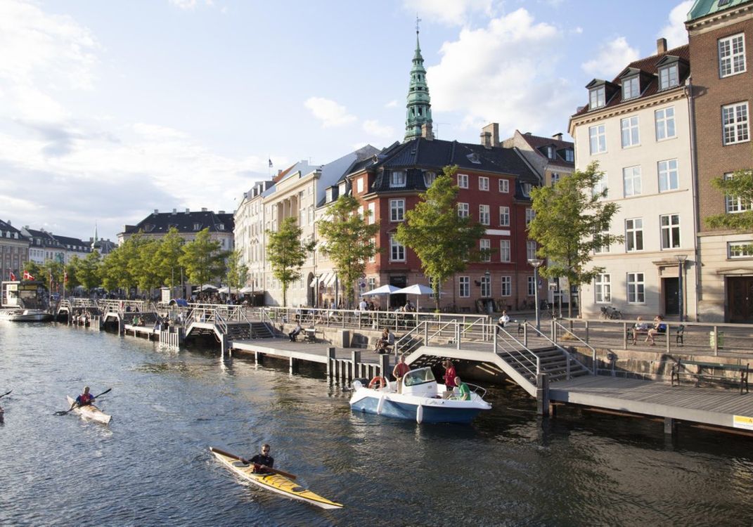 A canal in Denmark, specifically Københavns Kanaler, with kayakers and boaters on the water, lined by trees and buildings including a red brick structure with a green spire, under a partly cloudy sky.
