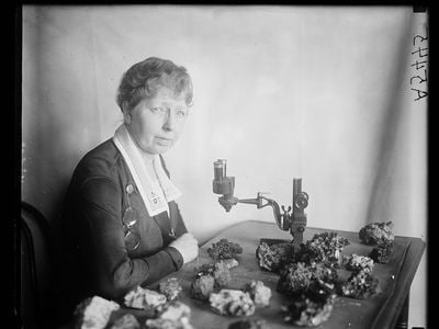Black and white photo of Margaret W. Moodey sitting at a desk with a microscope and gemstones