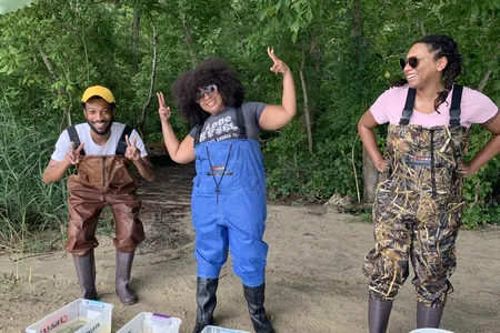 Three adults wearing chest waders laughing and in silly poses. They are standing on sand behind a line of plastic bins filled with water.