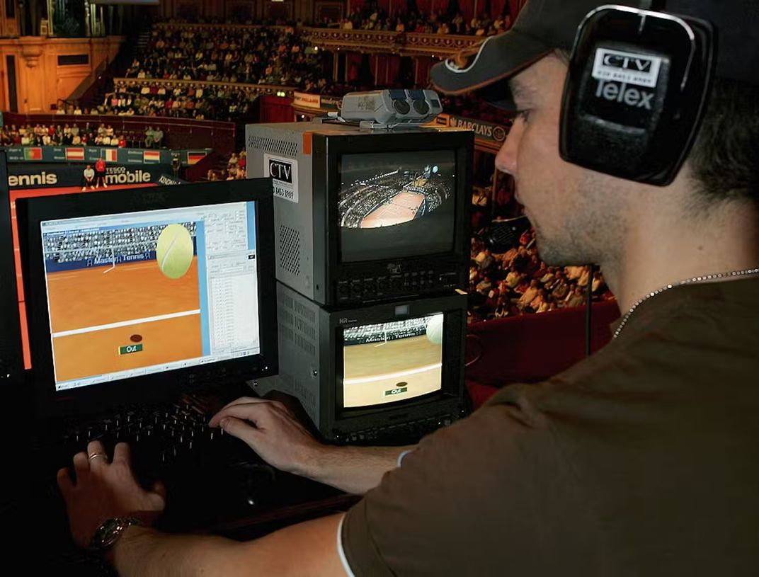 Technician wears headphones and works on a keyboard. He's looking at t a screen that shows the animation of a tennis ball.