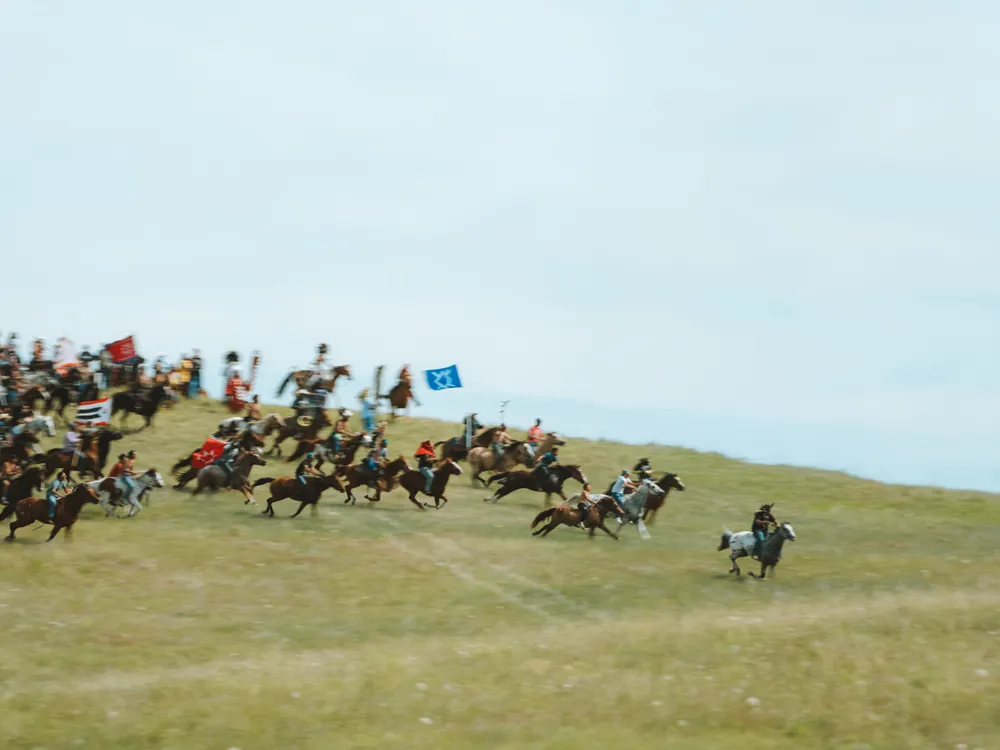 Dozens of riders on horseback race across an open grassland during a reenactment of the Battle of the Greasy Grass (Little Bighorn).