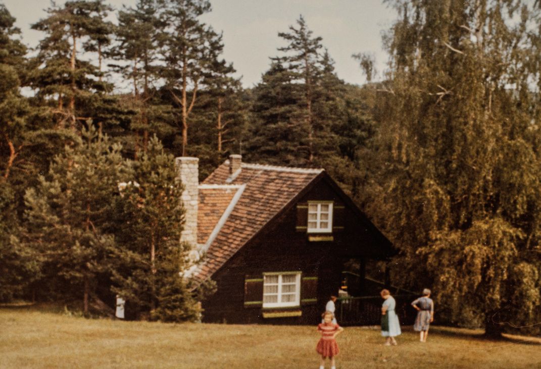 a group of people stand in front of a cottage in a wooded area on a hill