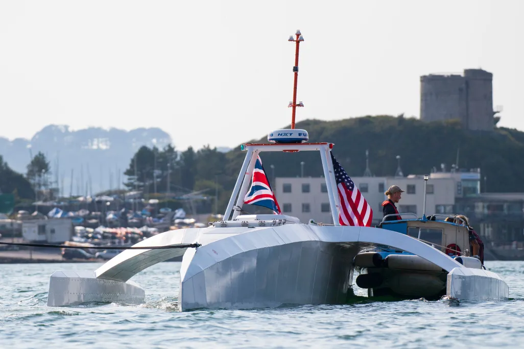 An oddly shaped metal ship with no windows, that floats on top of the water, with an American and British flag, is docked in a harbor
