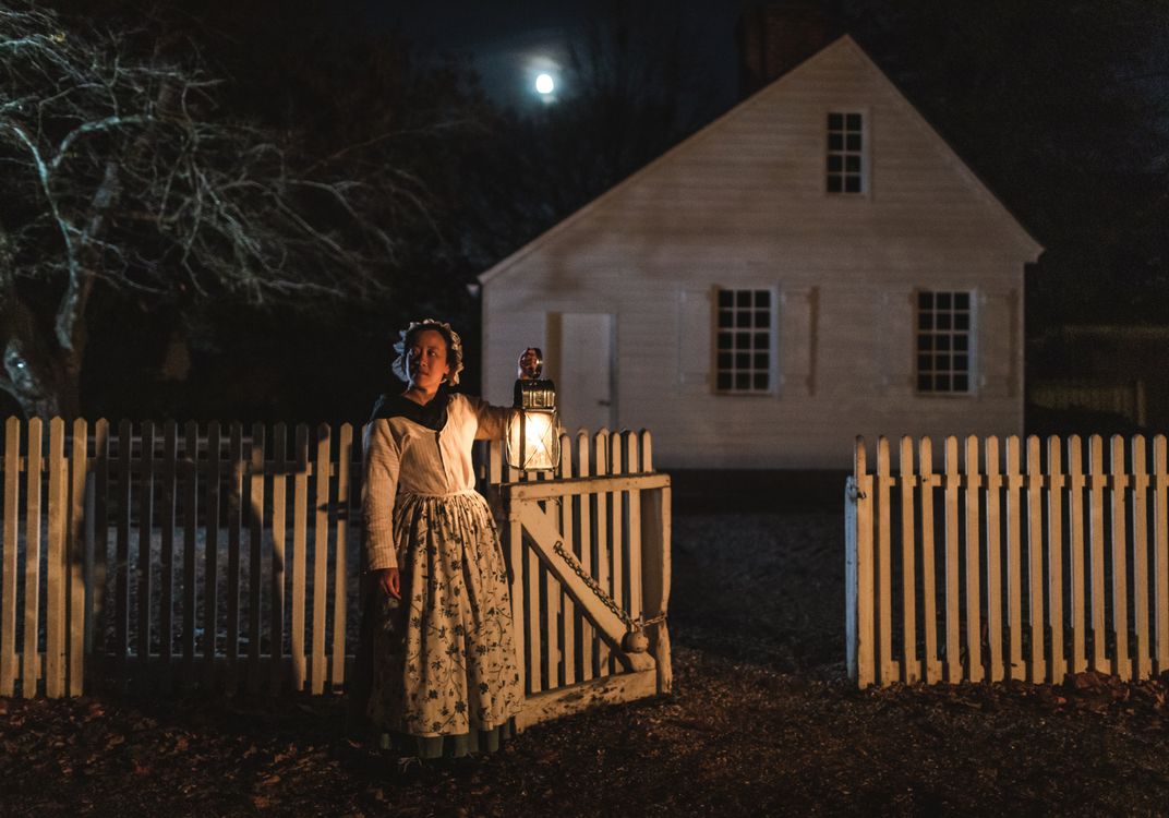 A person in period clothing stands at a white picket gate holding a lantern, illuminated against the darkness of night. Behind the fence, a historic white house sits beneath a bright moon, creating a scene reminiscent of a colonial-era evening.