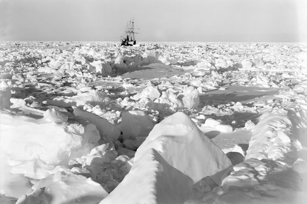 A view of a small ship leaning sideways in the background. In the foreground, a huge expanse of chunks of ice