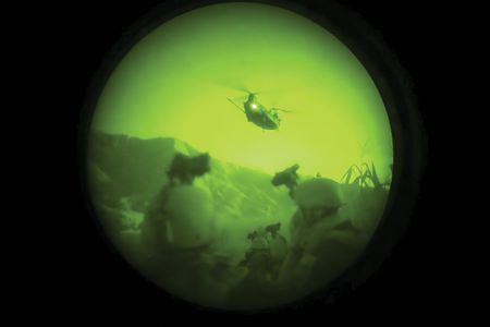 A grainy, green-tinted, circular view through a night vision scope shows a helicopter flying over a dark landscape at night.