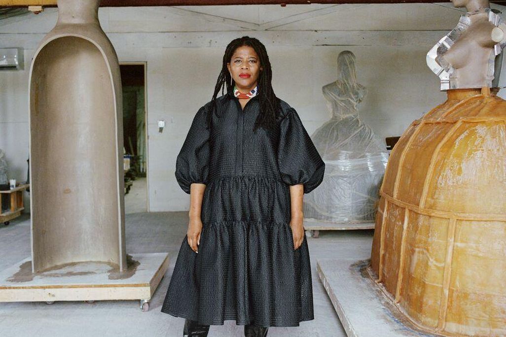 Simone Leigh, an African American woman, wears a voluminous black dress and stands in front of three of her sculptures, which are abstracted depictions of female figures on top of hoop skirts and jugs; Leigh faces the camera with a serious expression