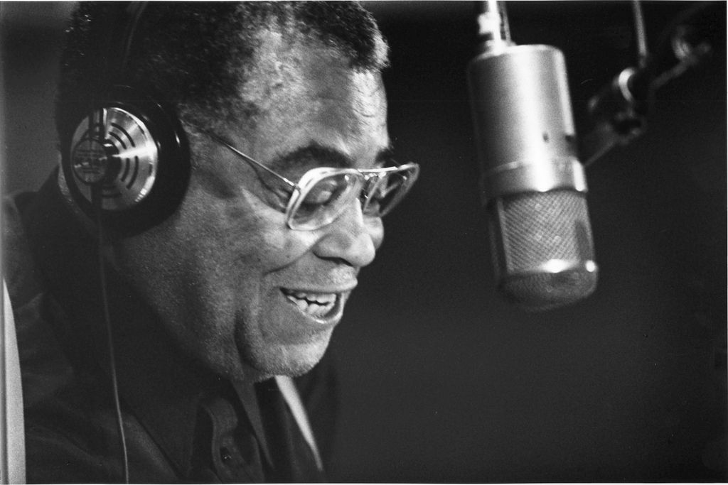 Black and white photo of a Black man wearing headphones and speaking into a microphone
