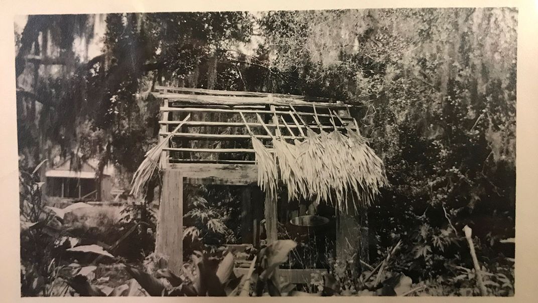The wooden frame of a small building with palm leaves forming an unfinished thatched roof. Black-and-white photo.