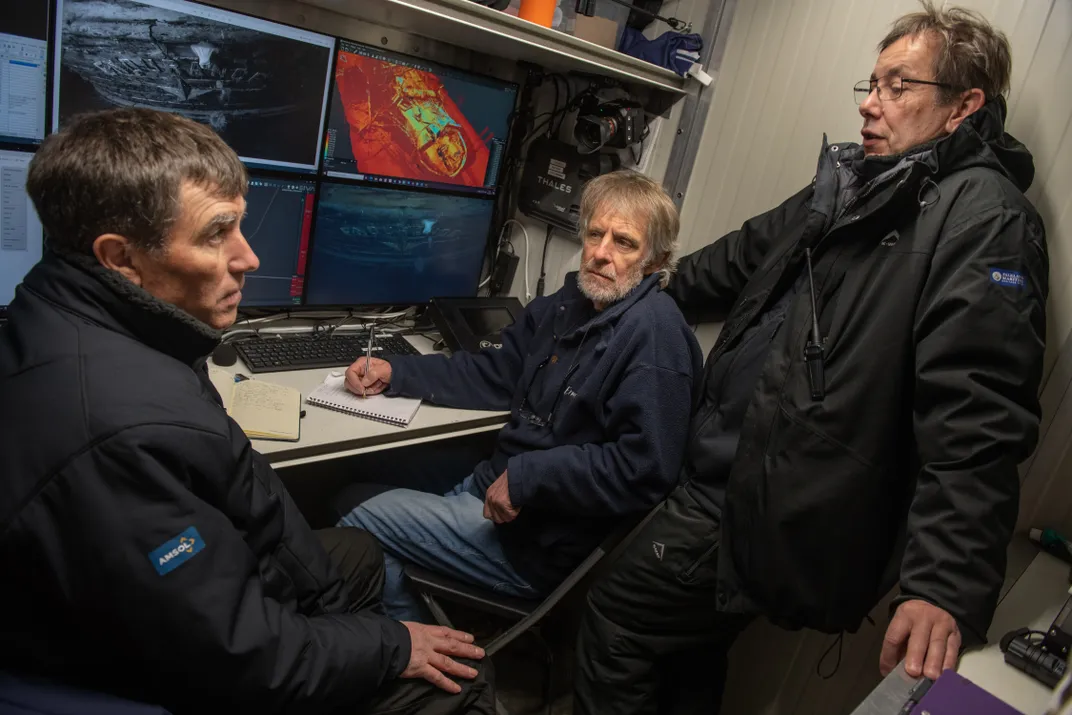 Three men sit around a set of computer screens showing infrared and color footage of the wreck