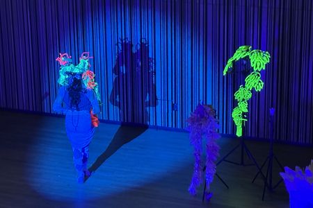 Armando Lopez-Bircann wears a cobalt blue bodysuit and skirt on a stage bathed in blue light. They have a neon green, blue, and pink headdress on and face away from the audience, with in front of their shadow. To the right are props including a neon green