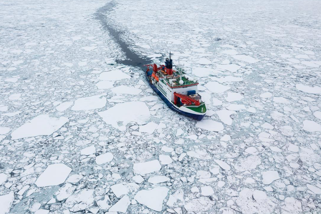 A photograph shows a white and red ship sailing through cracked sea ice 