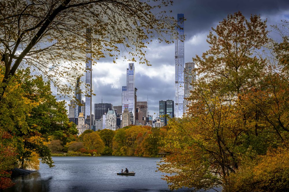 Autumn colors frame Central Park’s calm lake as two people row beneath looming skyscrapers. The contrast of vibrant foliage and dramatic city skyline captures a peaceful moment in the heart of New York.