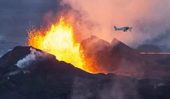 Red hot molten lava and thick dark dust spew from the ground while a small white. single-propeller aircraft flies overhead.