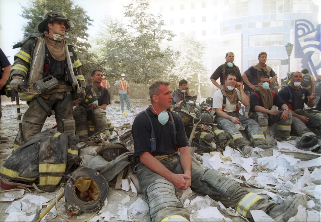 A group of men covered in white ash, looking exhausted, sit on the road and look out at the scene