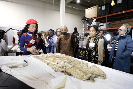 A group of people stand around a table looking at a dress laid out on top. Lighting and processing equipment is visible in the background.