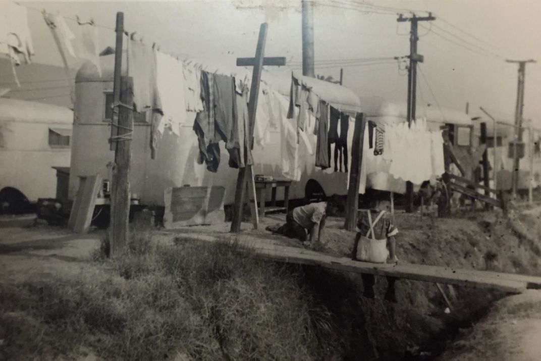 A black and white photo shows a child sitting below a clothesline in front of a row of white trailers.