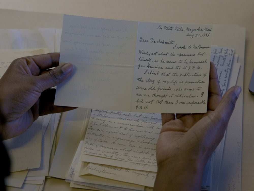 Two hands holding a card with handwritten information. A stack of other handwritten papers is visible on the table below.