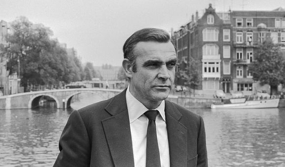 Image is black and white showing a man in a suit standing in front of a river