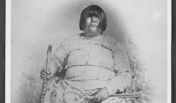 Black and white portrait of Dat So La Lee seated in a chair and surrounded by six decorative baskets on the floor by her feet.