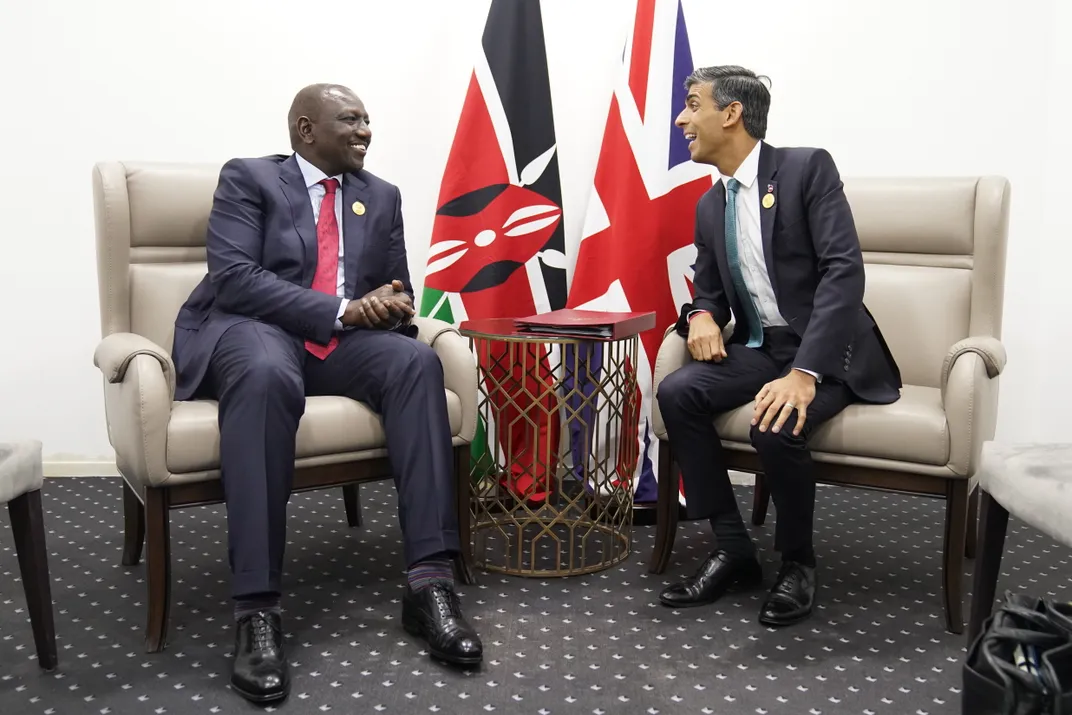two men talk and smile, sitting in front of their respective countries' flags