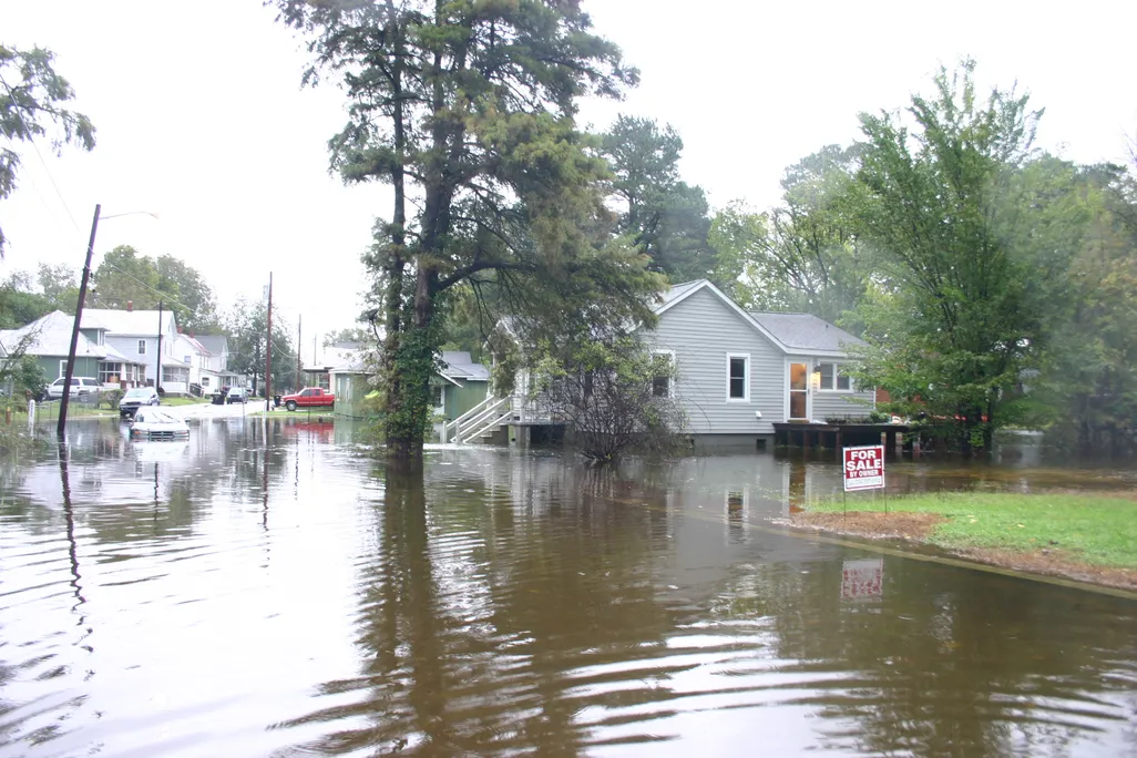 Image of a flooded neighborhood in North Carolina. The street is flooded with water and a car is halfway stuck in it. The water comes up to the houses' front porches. A "FOR SALE BY OWNER" sign is in a front yard.