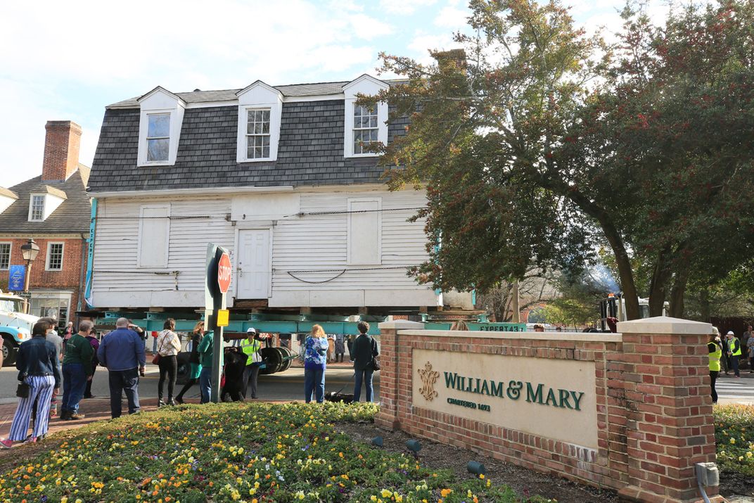 Large white house on truck bed in front of brick William & Mary sign