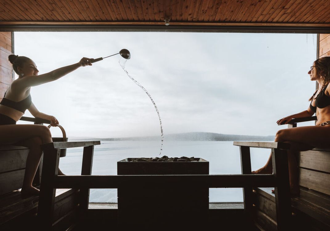 Two people in swimsuits sit in a sauna with a large window view of water; one pours water on hot stones to create steam.