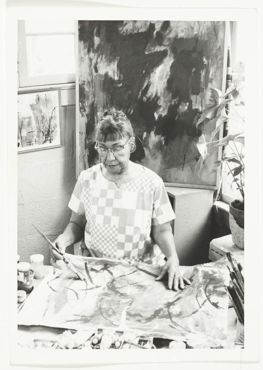 A black and white photograph of a woman in glasses painting on an unstretched canvas