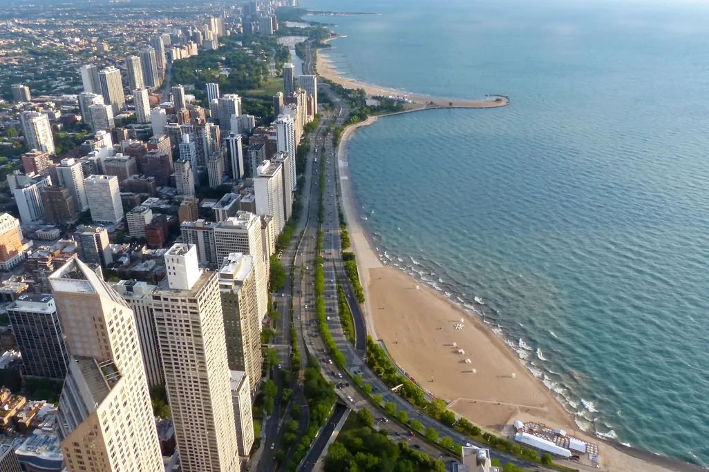A bright aerial view on a sunny day of Chicago, with skyscrapers on the left and the curve of a beach and a major two-lane highway in the middle, and blue, calm Lake Michigan on the right
