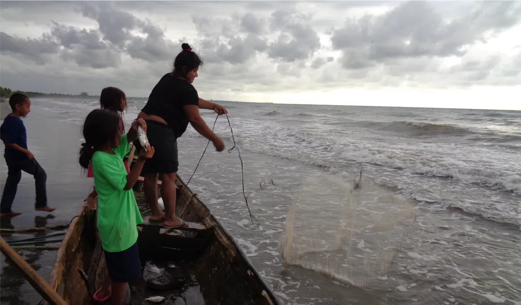 A woman and two children stand on a boat in shallow water while another child walks behind the boat. The woman holds a line attached to a fishing net over the boat, while the child in front holds a silver fish.