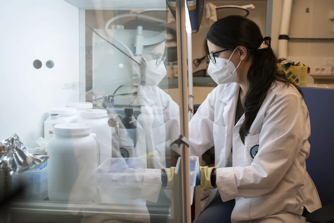 a female researcher in a lab coat, glasses and a mask sits in a chair in front of a glass case with materials for processing samples